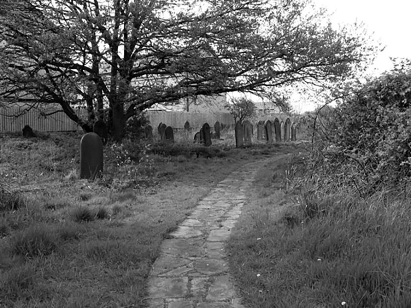Black and white image of Sculcoats cemetery with stone path, tree, and weathered gravestones under overcast sky